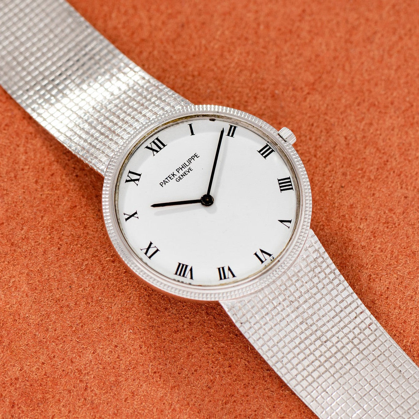 A silver Patek Philippe Calatrava watch with a white face and Roman numerals resting on an orange textured surface.