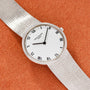 A silver Patek Philippe Calatrava watch with a white face and Roman numerals resting on an orange textured surface.