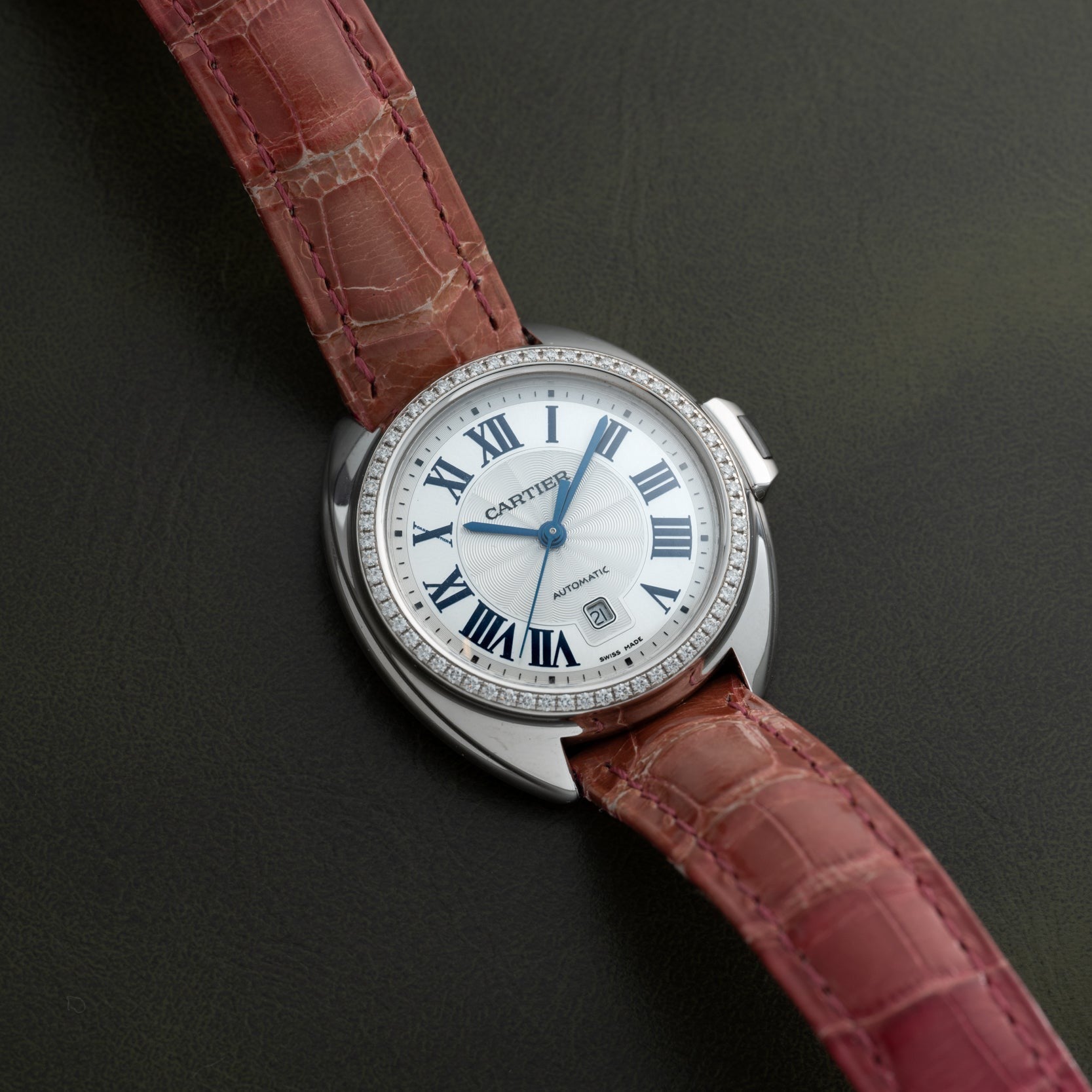 The Cartier Clé de Diamond watch, featuring a pink leather strap and diamond bezel, rests on a dark surface.