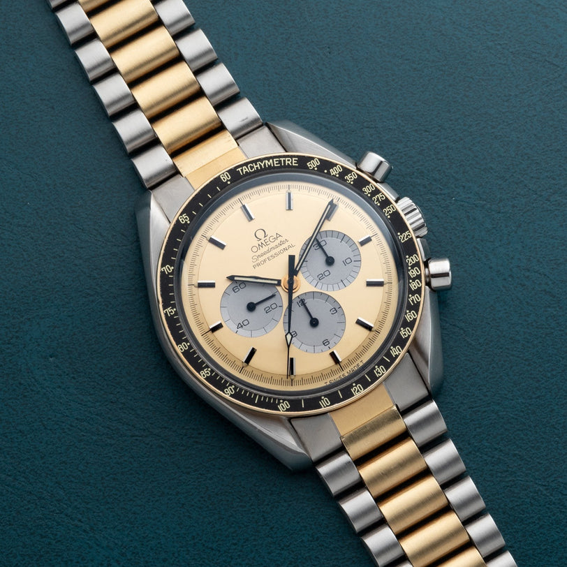 An OMEGA Speedmaster Professional Two-Tone watch, featuring gold and silver tones with a black bezel, displayed on a green leather surface.