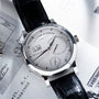 A Lange & Sohne Lange 31 wristwatch featuring a silver dial and black leather strap, displayed on a white paper background.