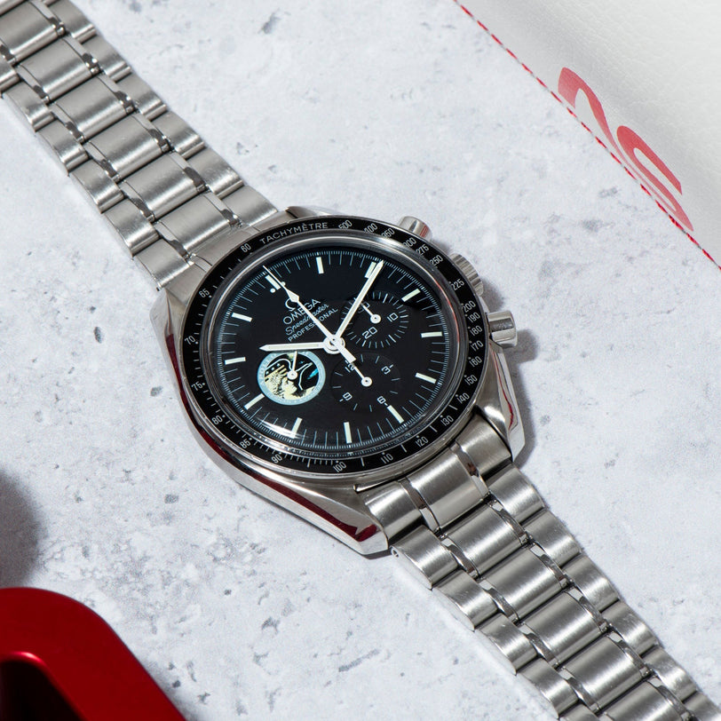 An OMEGA Speedmaster Professional Missions Apollo XVII wristwatch with a black dial and metal band rests on a light gray surface.