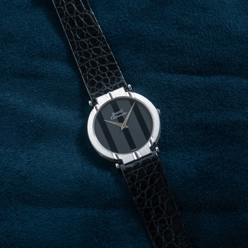 The Piaget White Gold Ladies luxury wristwatch with a black leather strap rests on a dark blue textured surface.