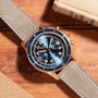 A Blancpain Air Command Flyback Chronograph with a blue face and grey strap rests on a wooden surface near two books.