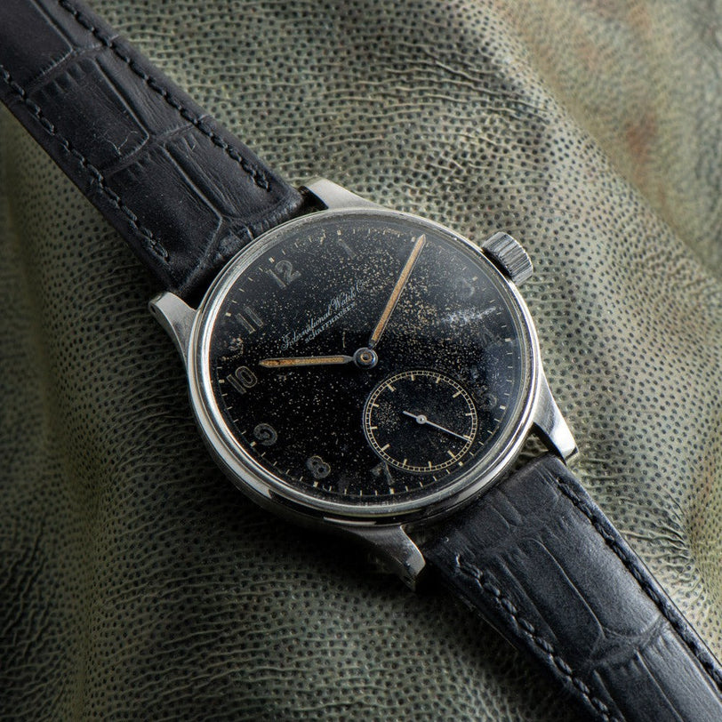 An IWC Schaffhausen Portugieser wristwatch with a worn black dial and black leather strap, displayed on textured green fabric.