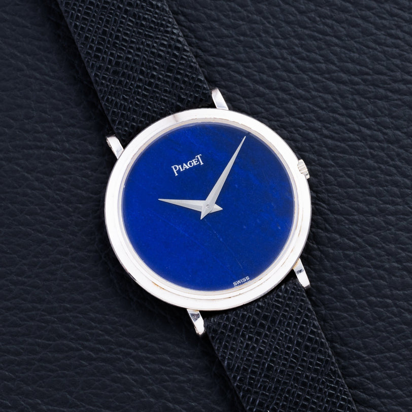 A Piaget Ladies Lapis Lazuli Dress Watch with a blue lapis face and black leather strap displayed on a textured black surface.