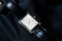 The Piaget Black Tie Emperador Coussin Automatique wristwatch, featuring a rectangular case and black leather strap, rests on dark fabric.