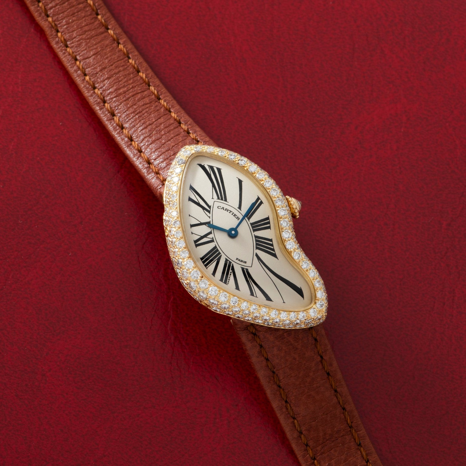 The Cartier Crash Diamonds is a luxury watch featuring Roman numerals, a diamond bezel, and a brown leather strap, shown on a red background.