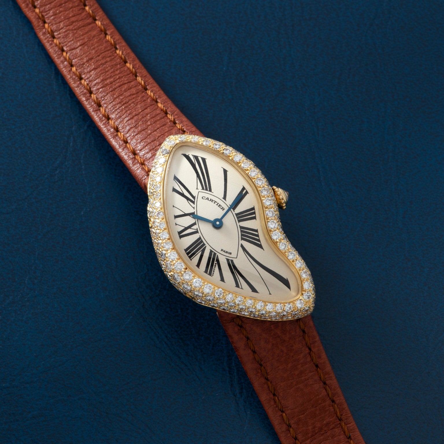 The Cartier Crash Diamonds watch features a brown leather strap, diamond bezel, and Roman numerals, showcased on a blue surface.