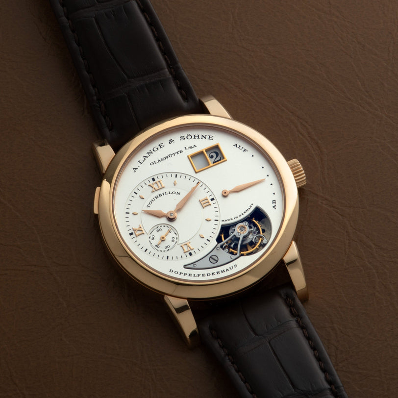 The Lange & Sohne Lange 1 Tourbillon luxury watch features a leather strap, gold case, and intricate dial, elegantly displayed on a brown surface.