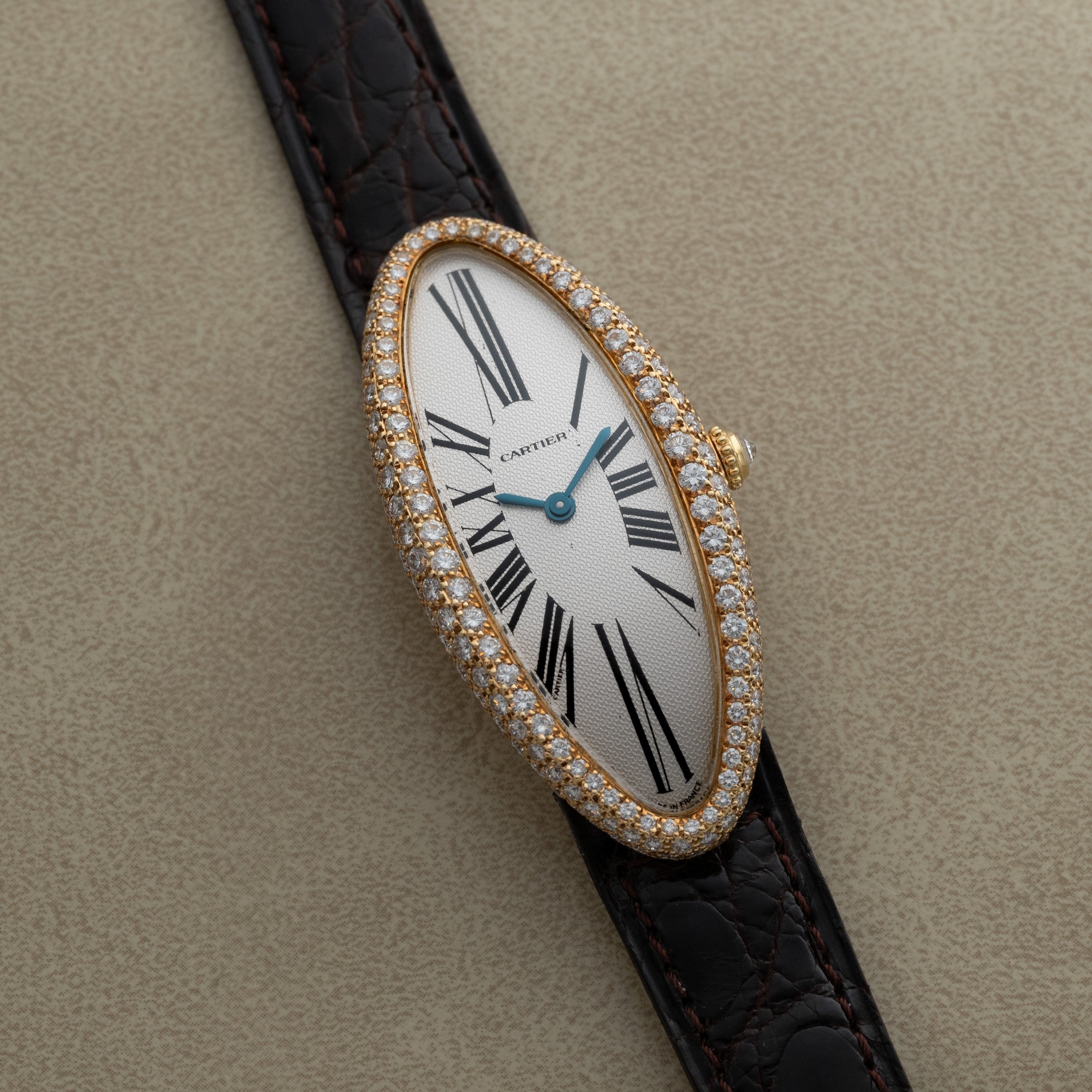 The Cartier Baignoire Allongee features an elongated oval face adorned with diamonds and a black leather strap, displayed on a beige surface.