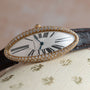 The Cartier Baignoire Allongee is an oval wristwatch with diamonds, Roman numerals, and a black leather strap.