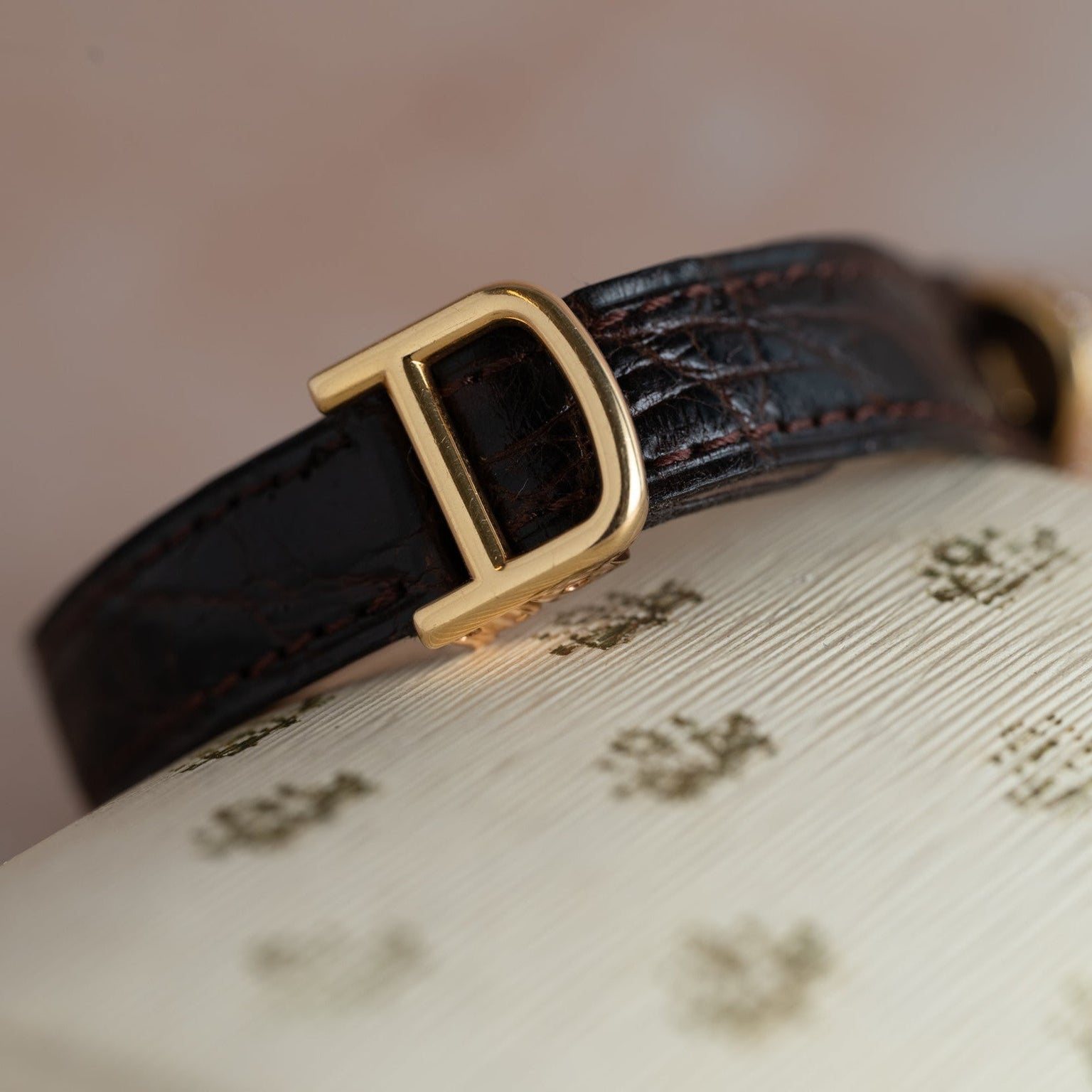 A Cartier Baignoire Allongee dark leather watch strap with a gold D buckle rests on a textured cream surface with gold patterns.