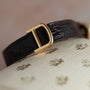 A Cartier Baignoire Allongee dark leather watch strap with a gold D buckle rests on a textured cream surface with gold patterns.