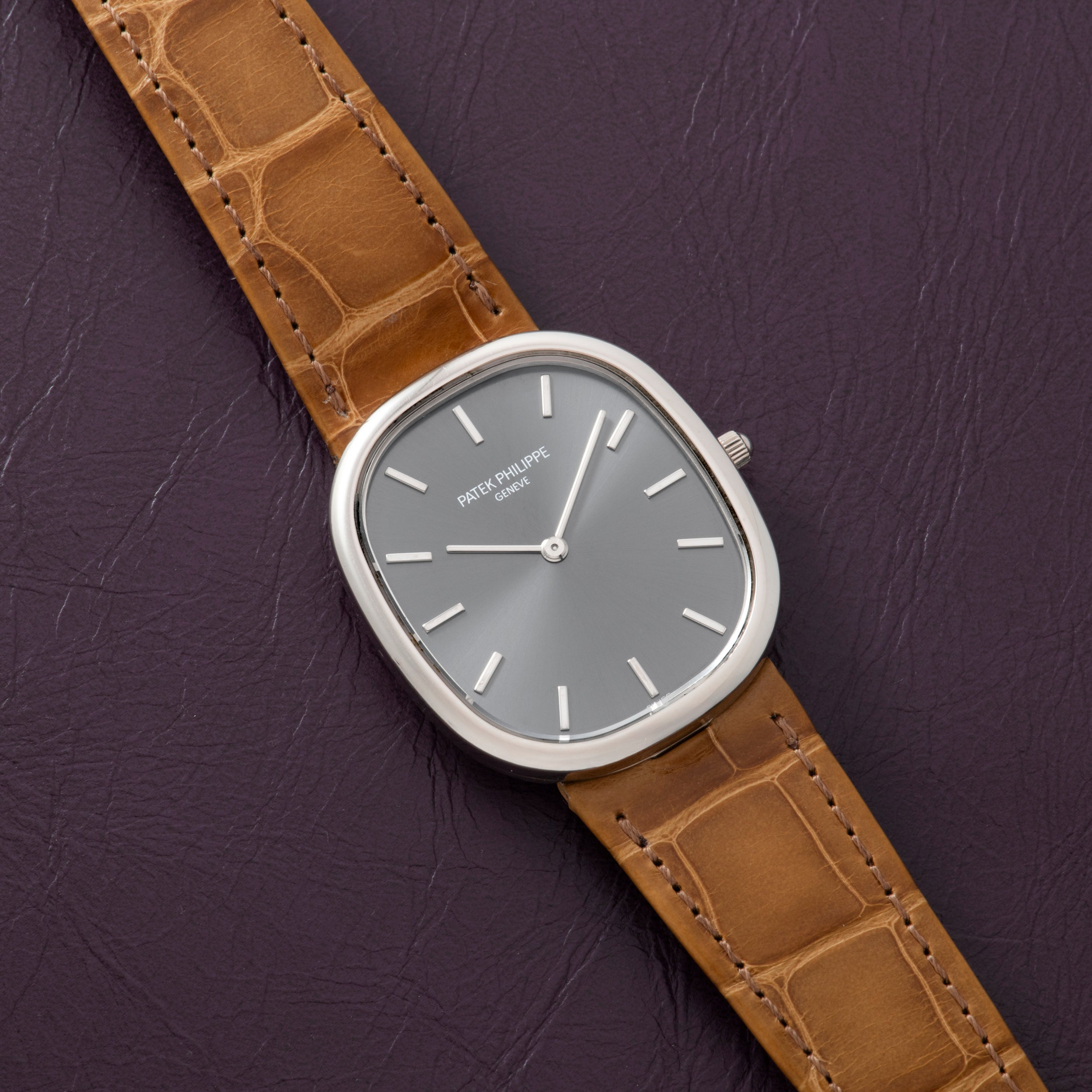 The Patek Philippe Ellipse watch with a brown leather strap and grey face is displayed on a textured dark purple surface.