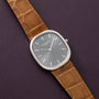 The Patek Philippe Ellipse watch with a brown leather strap and grey face is displayed on a textured dark purple surface.