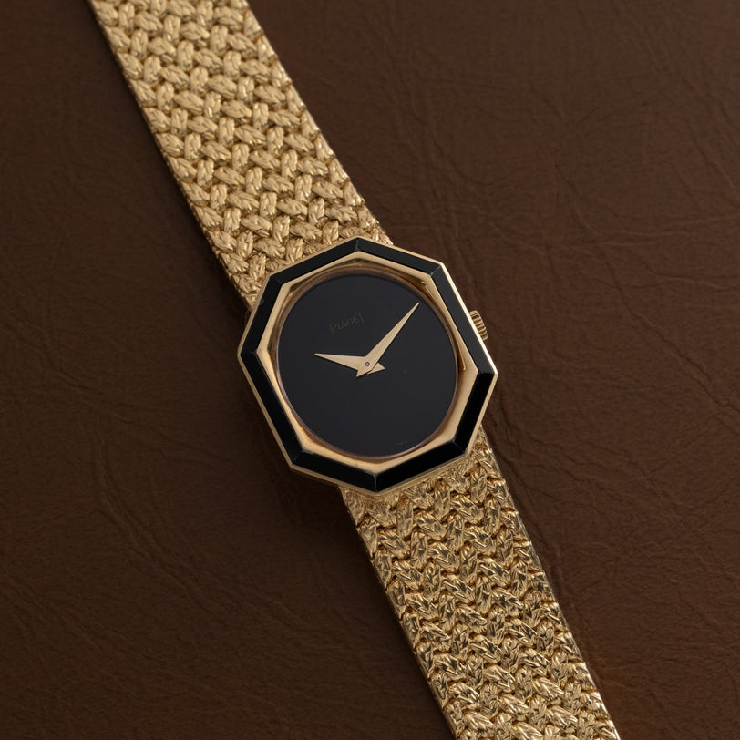 A Piaget Onyx Dial gold watch with a black octagonal face and woven gold band rests on a brown leather surface.