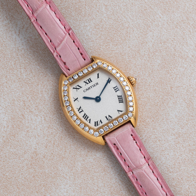 The Cartier Tortue watch features a pink leather strap, diamond bezel, and Roman numeral dial, displayed on a beige surface.