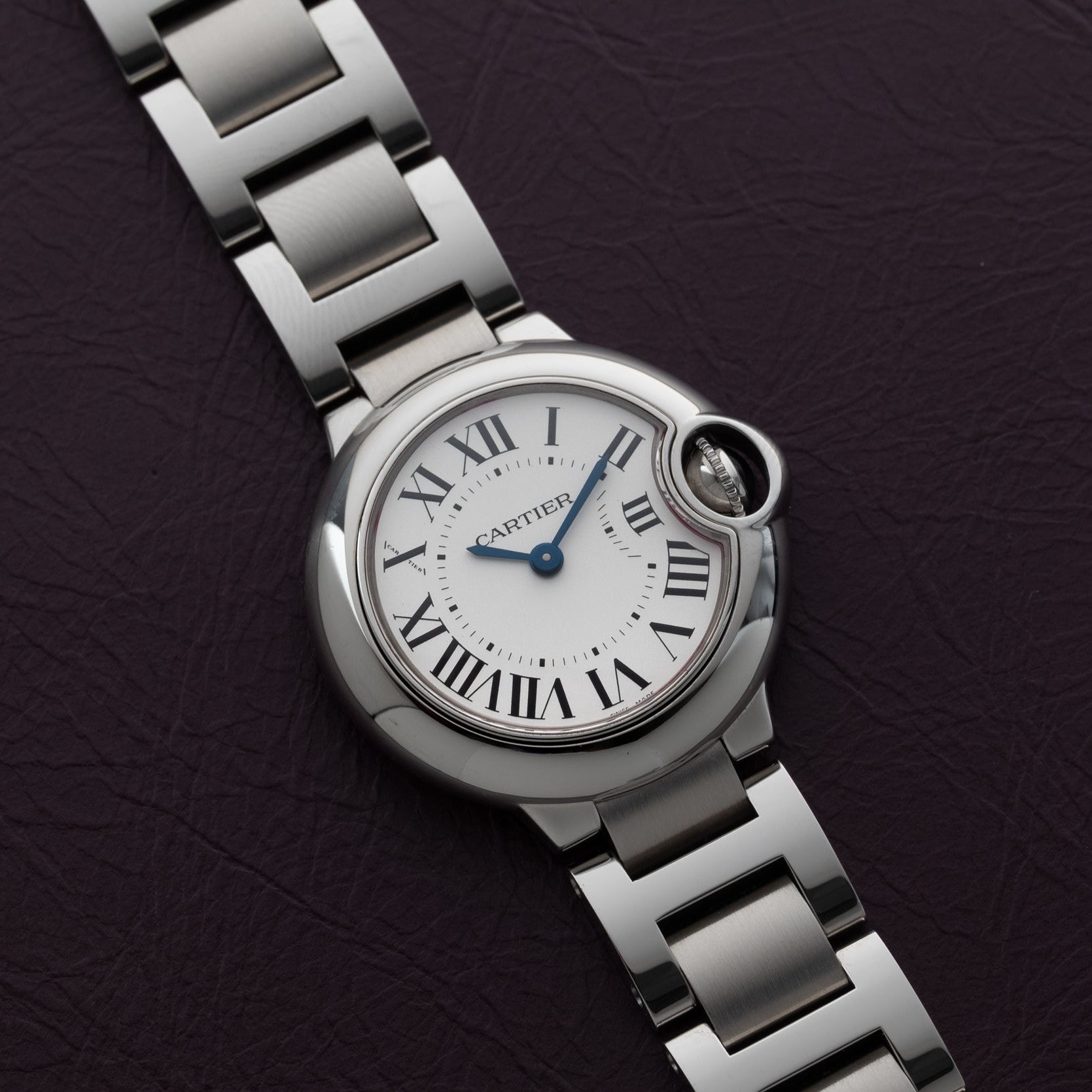 A Cartier Ballon Bleu silver wristwatch with Roman numerals rests on a dark textured surface.