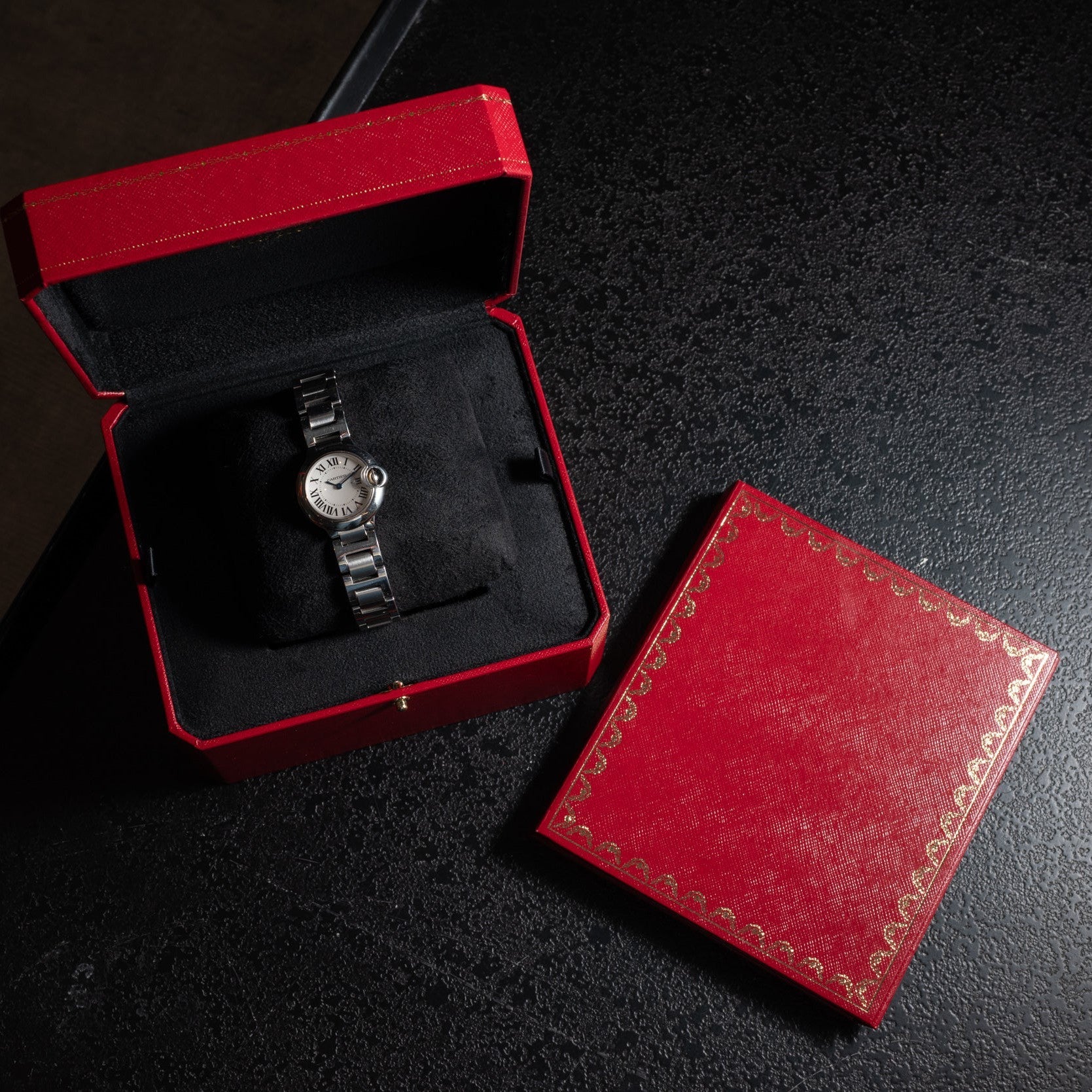 A Cartier Ballon Bleu silver wristwatch is displayed on a dark surface, presented in a red box with a matching red lid.