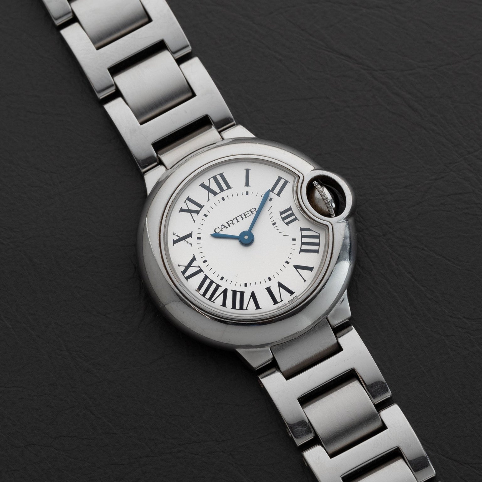 The Ballon Bleu by Cartier is a silver wristwatch featuring Roman numerals on a black leather strap.