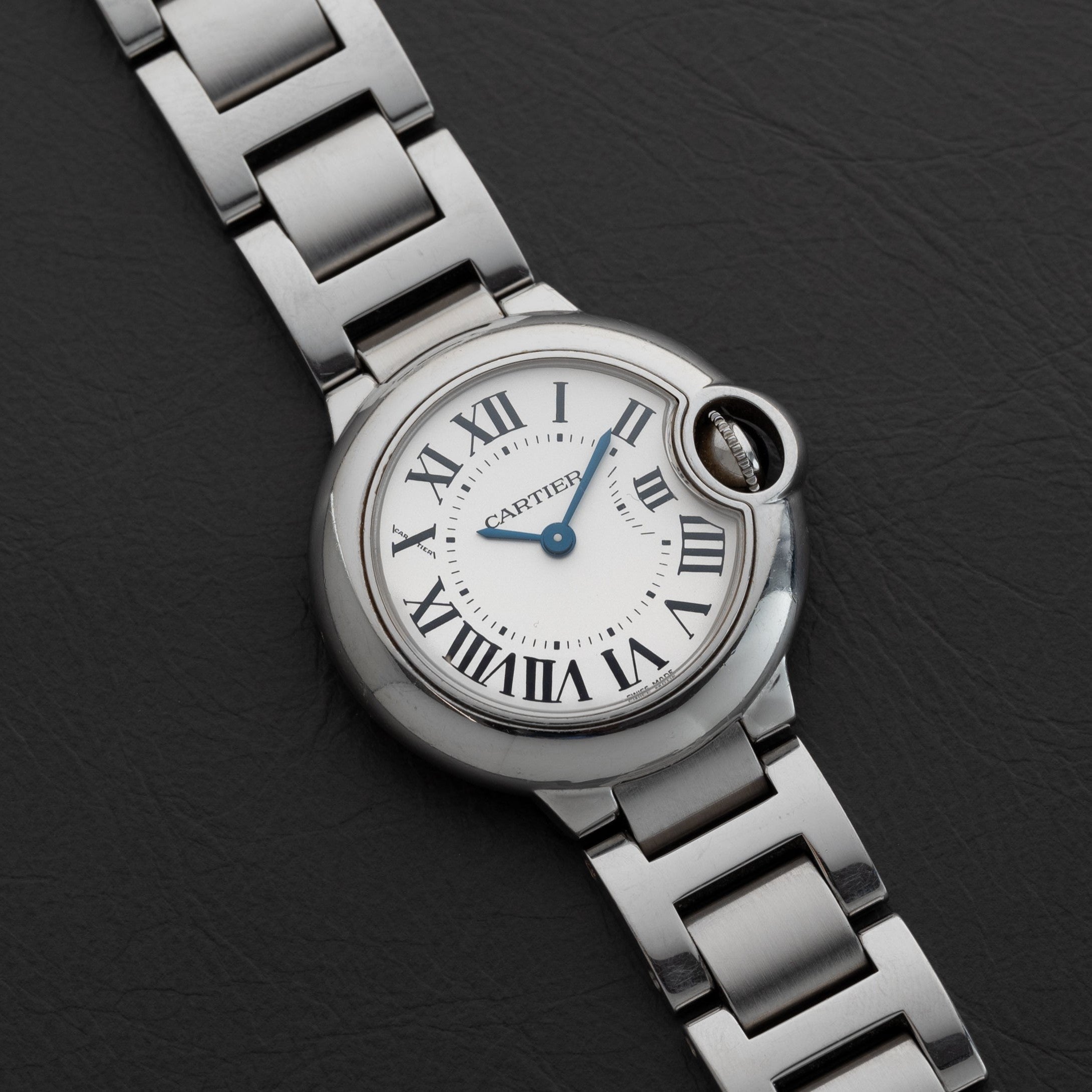 The Ballon Bleu by Cartier is a silver wristwatch featuring Roman numerals on a black leather strap.