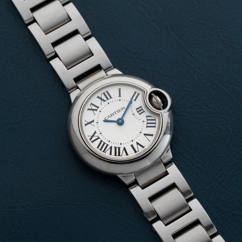 The Cartier Ballon Bleu wristwatch, featuring Roman numerals, rests on a dark textured surface displaying the time 10:11.