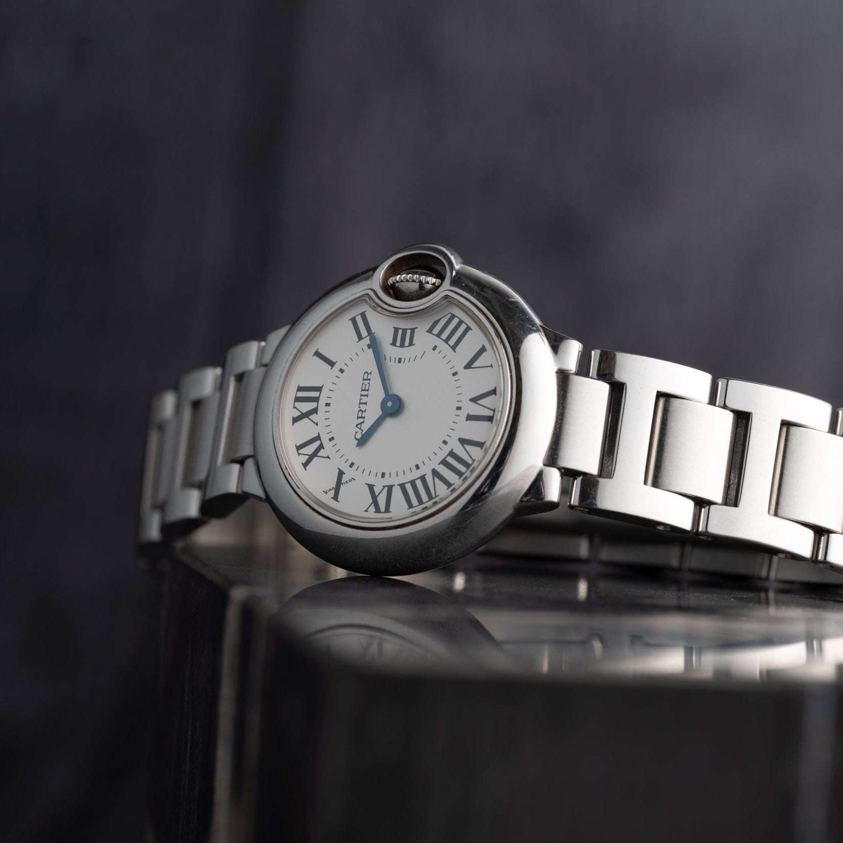 The Cartier Ballon Bleu, a silver wristwatch with Roman numerals and a white dial, rests on a reflective surface.