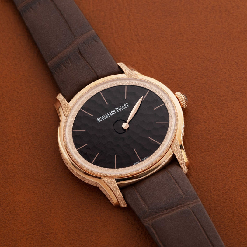 The Audemars Piguet Millenary Philosophique features a gold case, black textured dial, and brown leather strap, displayed against a brown background.