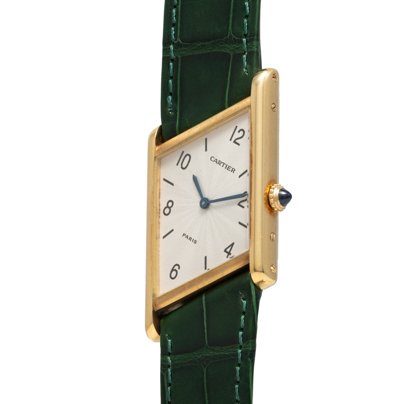 The Cartier Tank Asymétrique wristwatch features a gold case, white face, blue hands, and a green leather strap, shown at an angle.