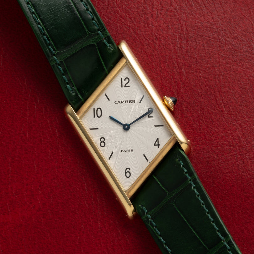 A Cartier Tank Asymétrique gold watch with a green leather strap rests on a red leather surface.