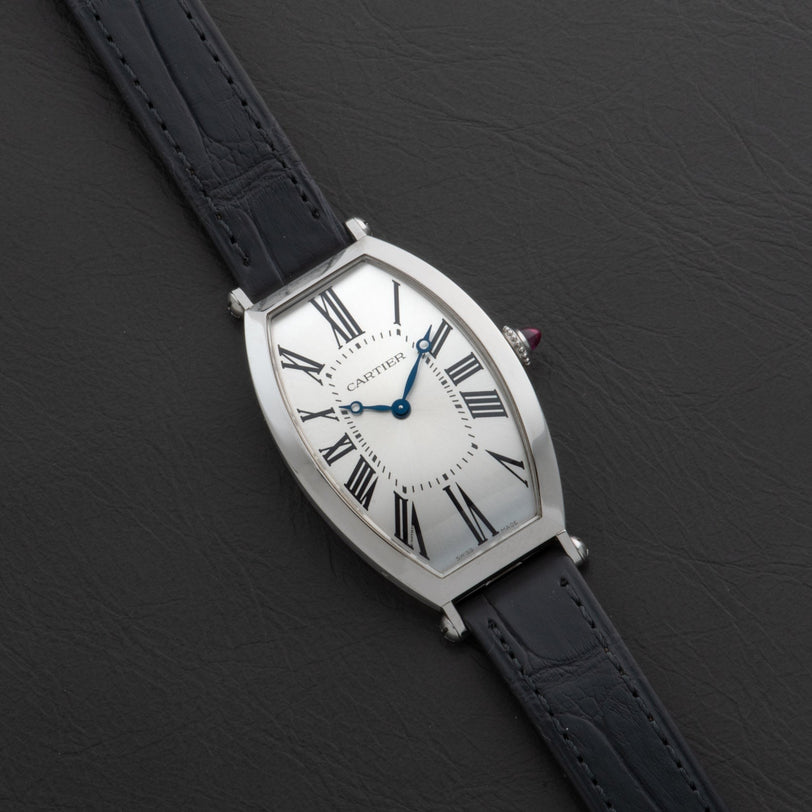 The Cartier Tonneau Platinum watch features a white face, Roman numerals, and a black leather strap, displayed on a dark surface.