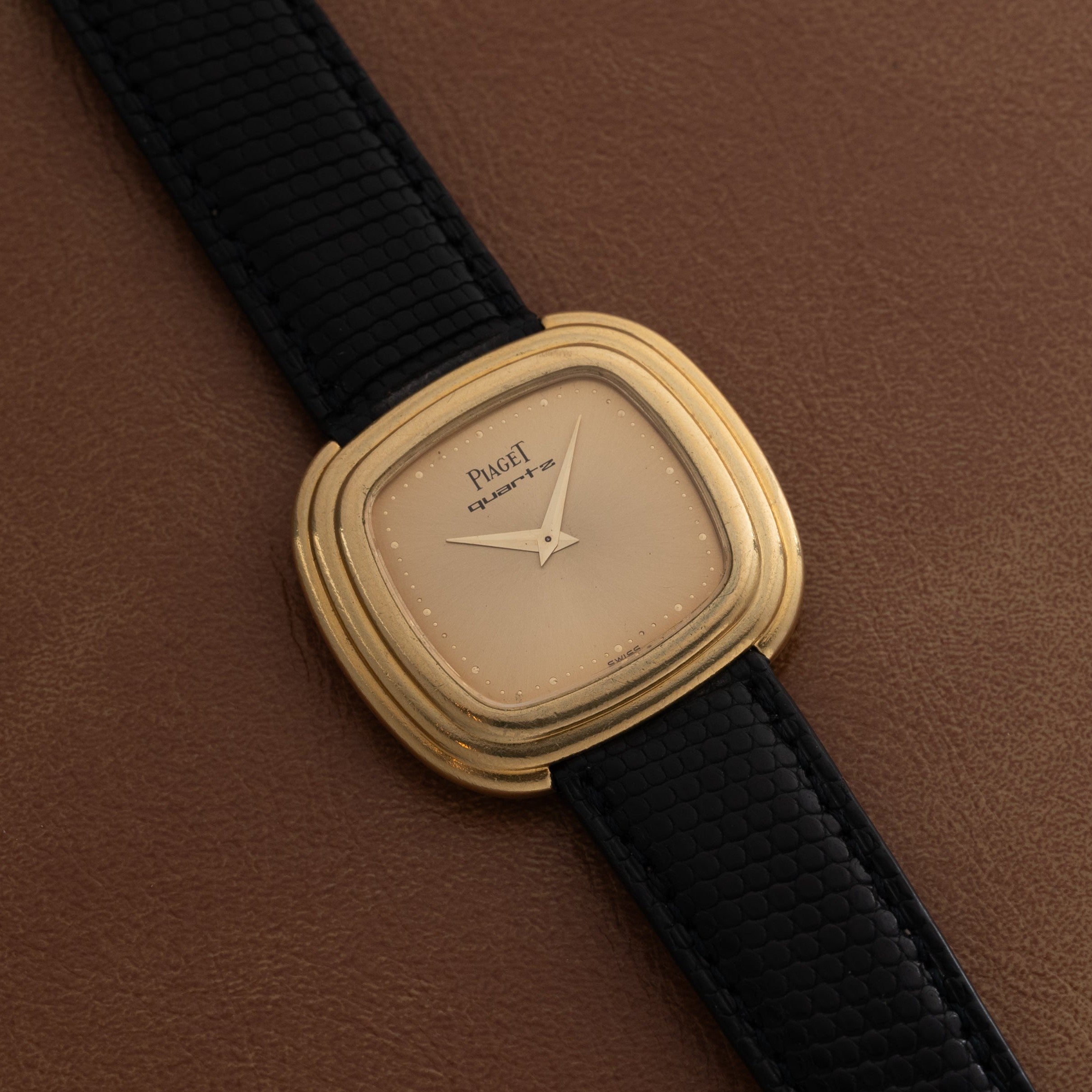 A Piaget Emperador Mini Warhol gold wristwatch with a square face and black leather strap rests on a brown surface.