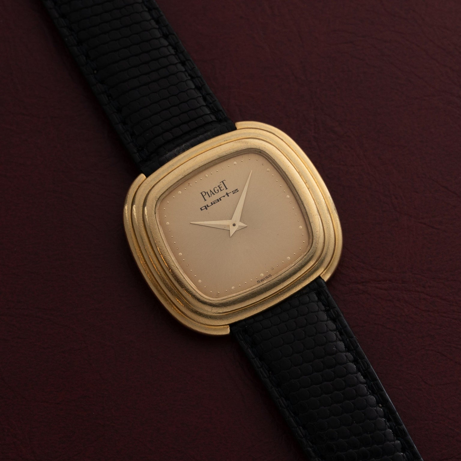 A Piaget Emperador Mini Warhol gold wristwatch with a black textured leather strap displayed on a dark maroon surface.