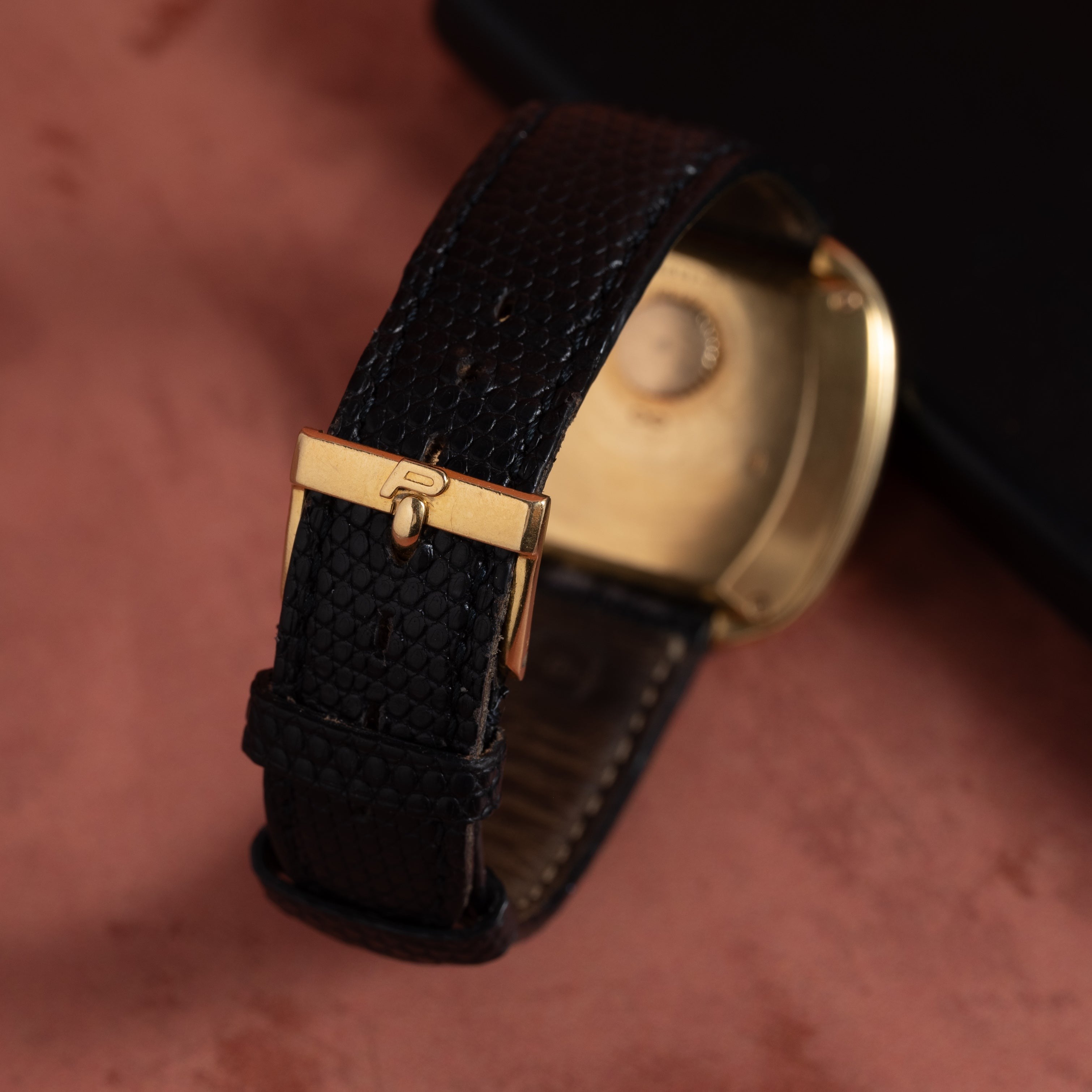 Close-up of the Piaget Emperador Mini Warhol gold watch featuring a black textured leather strap and gold buckle, displayed on a reddish surface.