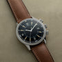 The Jaeger-LeCoultre Tribute to Polaris, a vintage-style watch with a brown leather strap and black dial, rests on a beige surface.