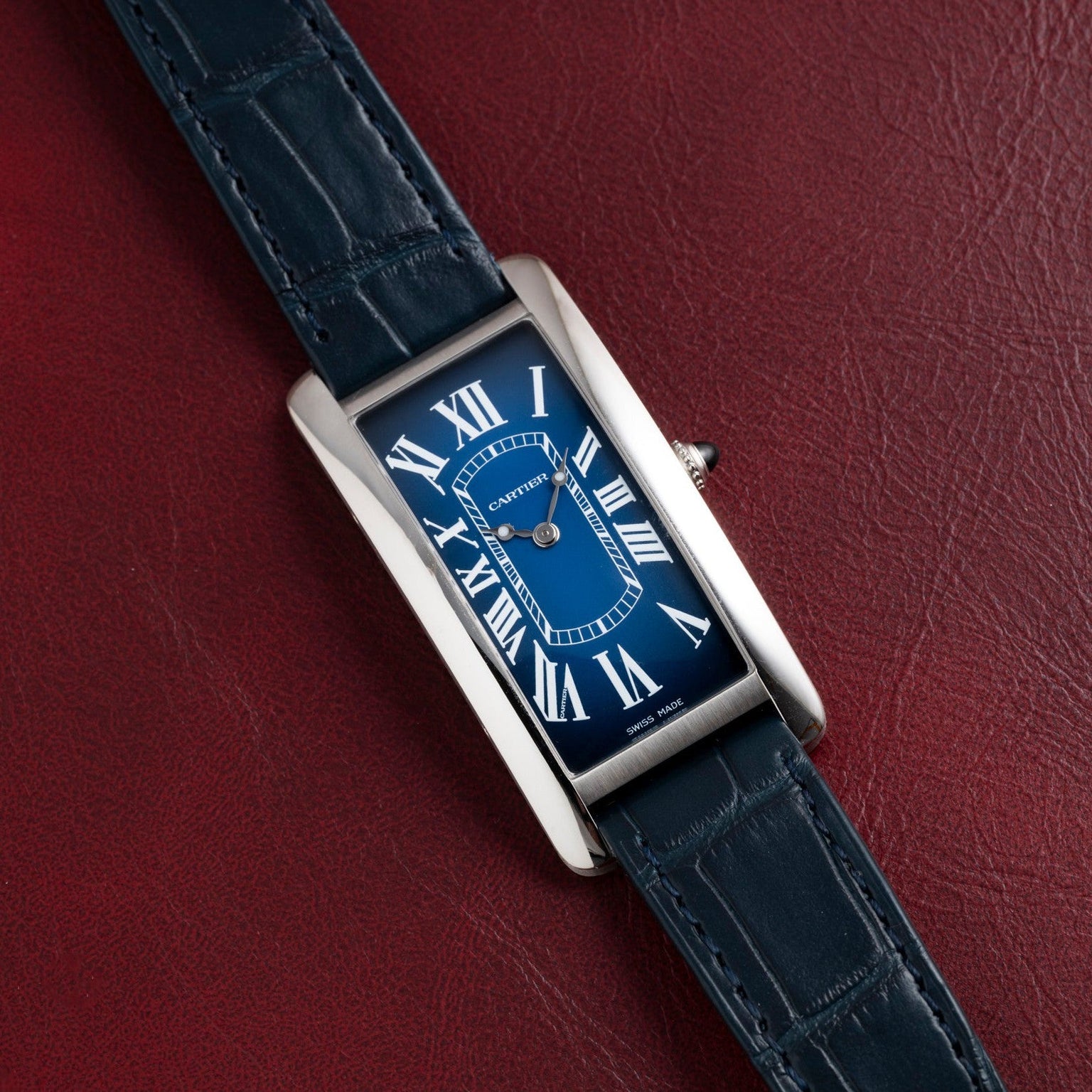 A blue Cartier Tank Cintrée watch with Roman numerals and a dark blue leather strap rests on red leather.