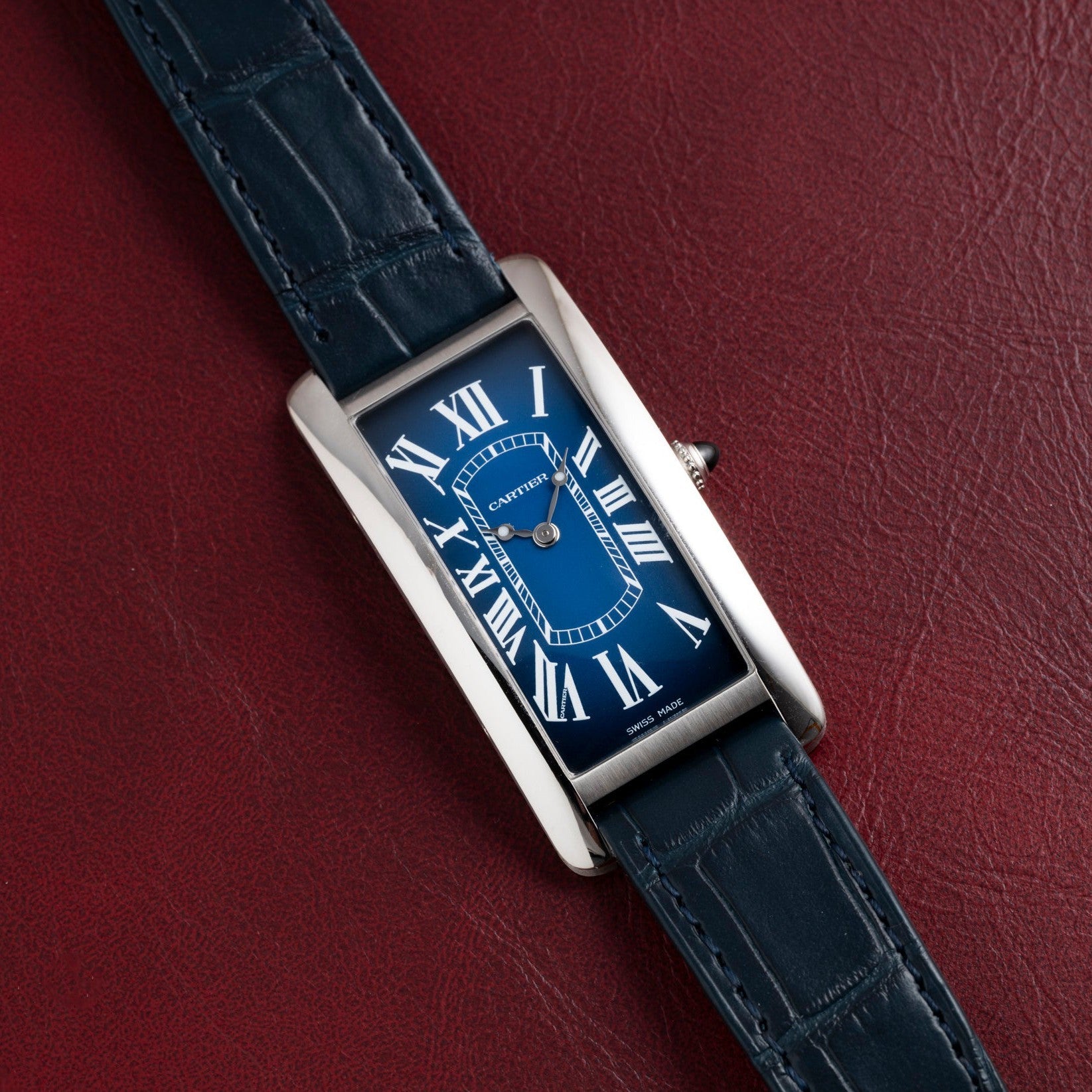 A blue Cartier Tank Cintrée watch with Roman numerals and a dark blue leather strap rests on red leather.