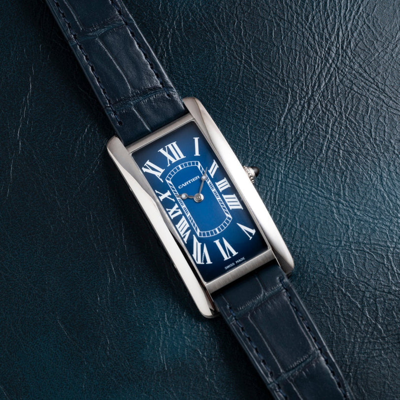 A Cartier Tank Cintrée wristwatch with a blue leather strap and Roman numerals on a textured dark surface.