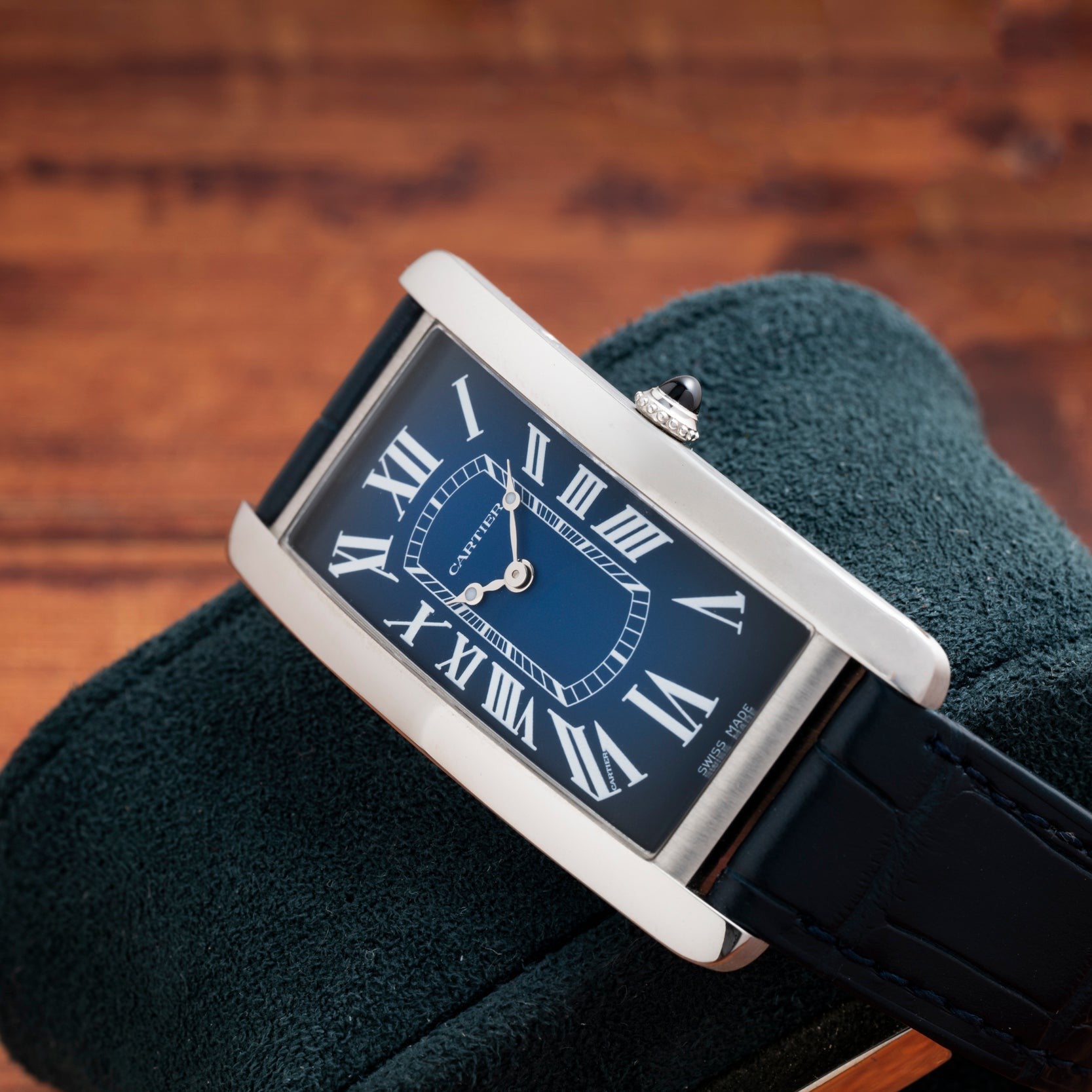 The Cartier Tank Cintrée, a rectangular blue watch with Roman numerals, rests on a dark green suede cushion against a wooden background.