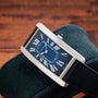 The Cartier Tank Cintrée, a rectangular blue watch with Roman numerals, rests on a dark green suede cushion against a wooden background.