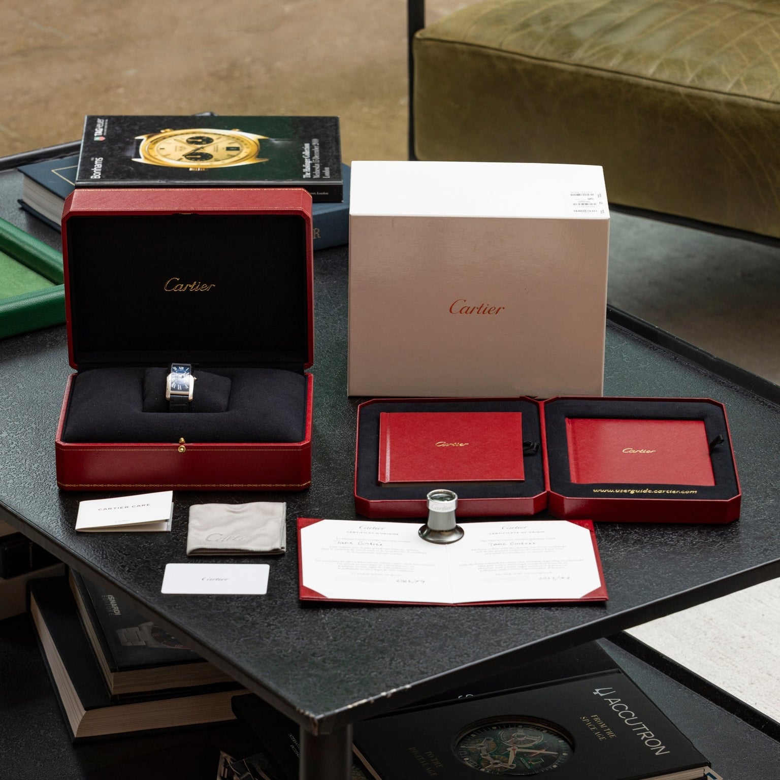 The Cartier Tank Cintrée watch, complete with its red box, papers, loupe, and packaging, is displayed on a black table.