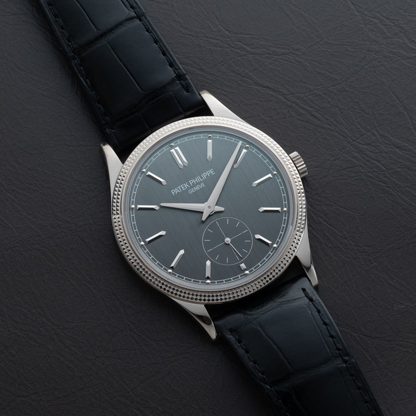 A Patek Philippe Calatrava wristwatch with a silver case and black leather strap displayed on a dark leather surface.