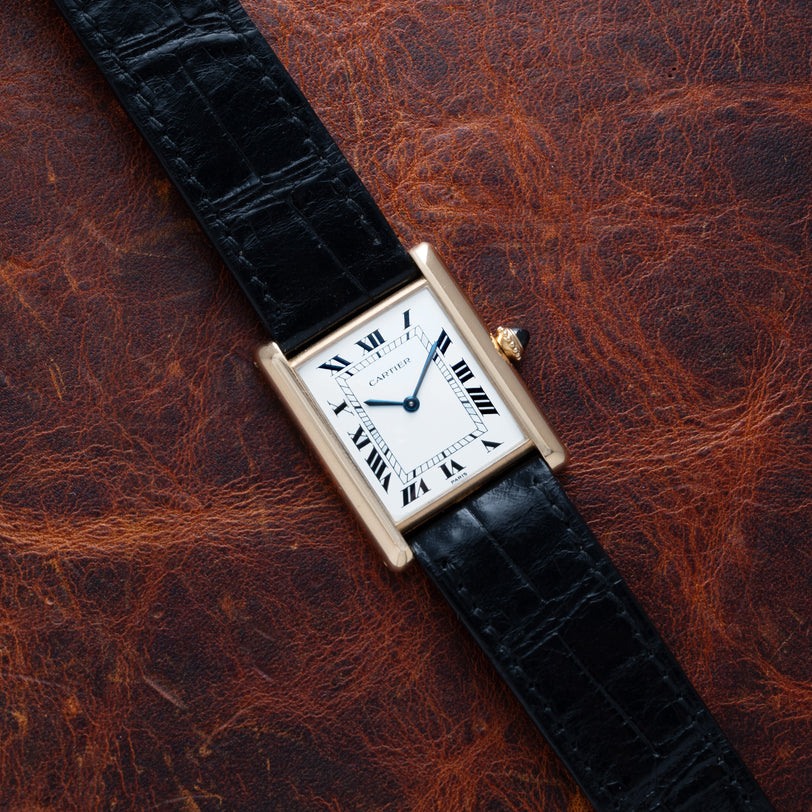 A Cartier Tank Louis watch with a black leather strap rests on a textured brown leather surface.