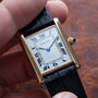 A hand holding a Cartier Tank Louis watch featuring a gold case, Roman numerals, and a black leather strap.