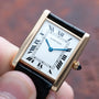 A hand holding a Cartier Tank Louis watch with a square face, Roman numerals, and a black leather strap.