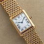 A Cartier Tank Louis gold wristwatch with a rectangular face, Roman numerals, and mesh bracelet rests on a beige surface.