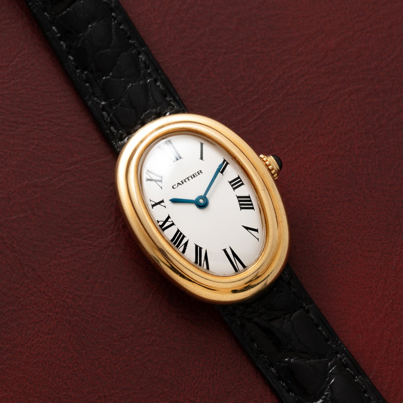 Cartier Baignoire wristwatch with Roman numerals and a black leather strap, displayed on a dark red leather surface.