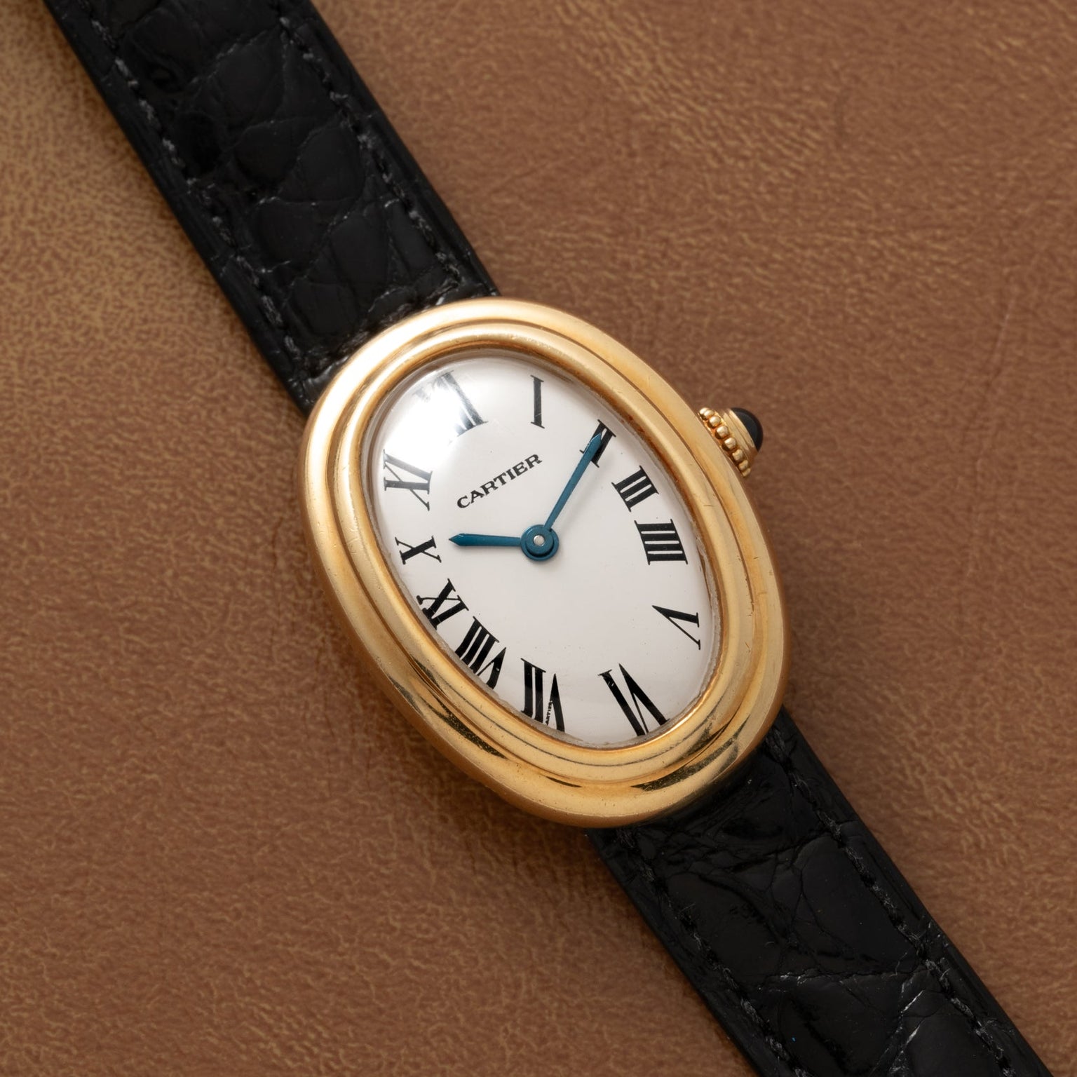 A Cartier Baignoire wristwatch with a white oval face, Roman numerals, and a black leather strap on brown leather.