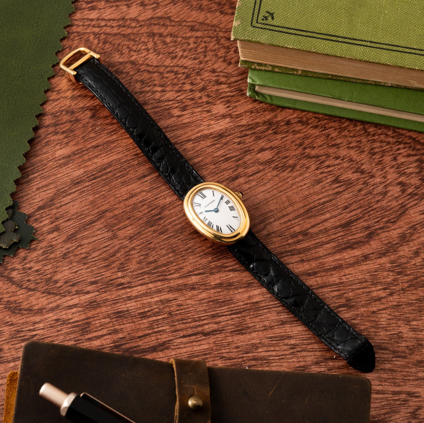 The Cartier Baignoire gold watch with a black leather strap rests on a wooden desk next to green books and a brown notebook.